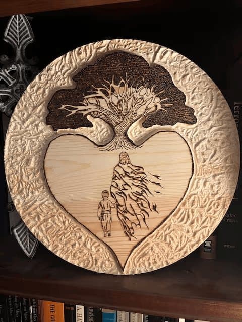 A round wood carving of a tree above a heart, with a child and a robed figure walking together inside the heart.