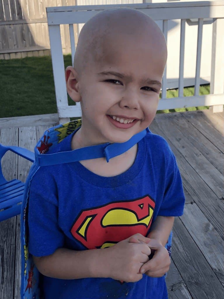 Jacob stands on a wooden deck in a Superman shirt and blue cape, hands clasped, smiling at the camera.