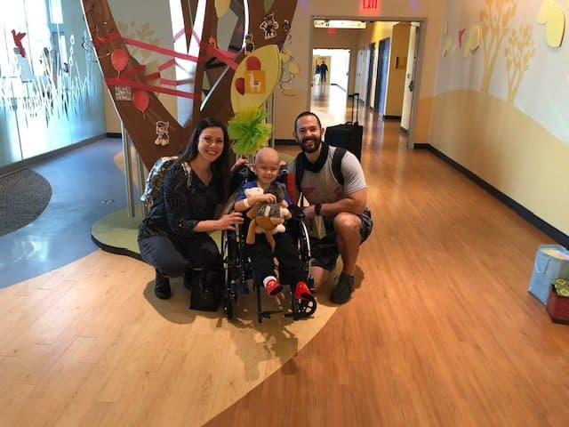 Jacob sits in a wheelchair holding a stuffed animal while his mom and dad kneel on either side of him, all three smiling in a bright hallway.