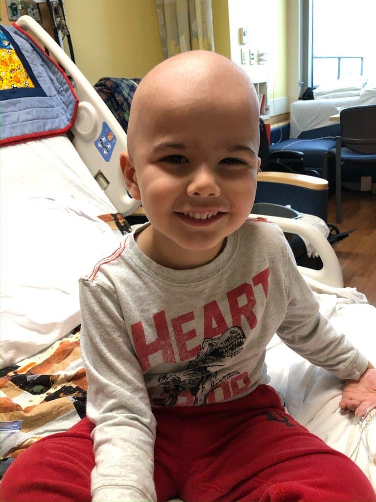 Jacob sits on a hospital bed in a gray shirt with a red heart graphic and smiles at the camera.