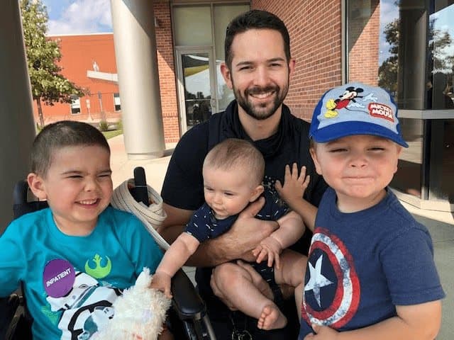 Jacob sits with a stuffed animal beside his dad, who is holding a baby, while a brother in a Mickey Mouse cap stands with them outside a brick building.