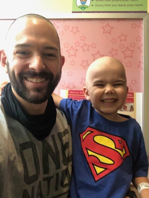 Jacob in a Superman shirt laughs with his eyes squeezed shut while his dad smiles beside him.