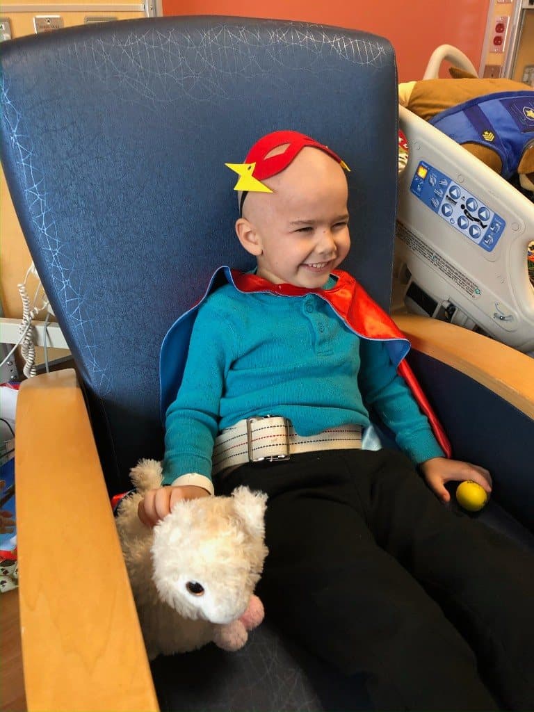 Jacob grins in a big blue chair wearing a handmade red mask and cape, holding a stuffed dog, with a yellow ball beside him.